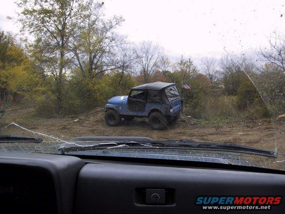 Blue Jeep Wrangler with soft top on a dirt path, viewed from inside another vehicle