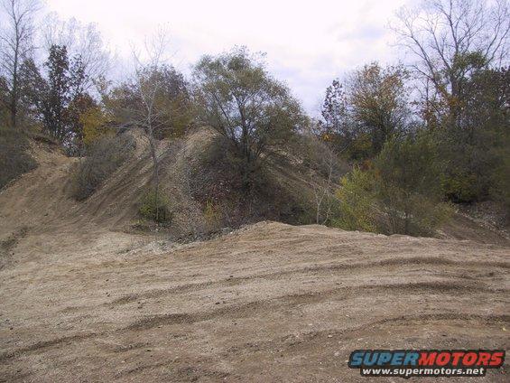 Dirt hill with tire tracks and trees under a cloudy sky. Supermotors.net logo in lower right