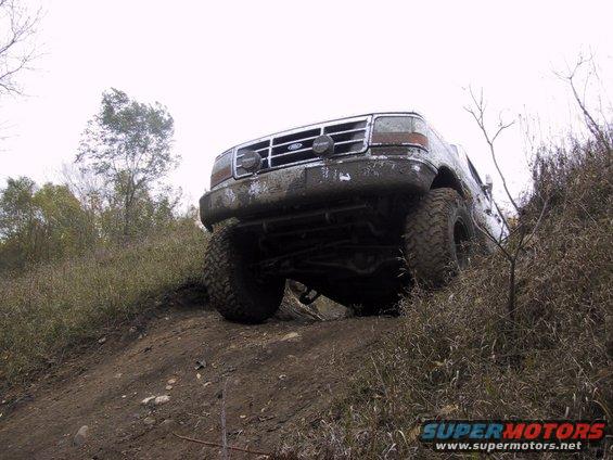 Mud-covered white Ford Bronco driving up a dirt hill, front 3/4 view