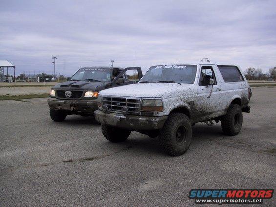 White Ford Bronco and black Ford F-150 covered in mud, parked on pavement