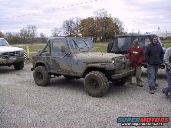 Muddy Jeep Wrangler parked on gravel with people around. Another truck is to the left