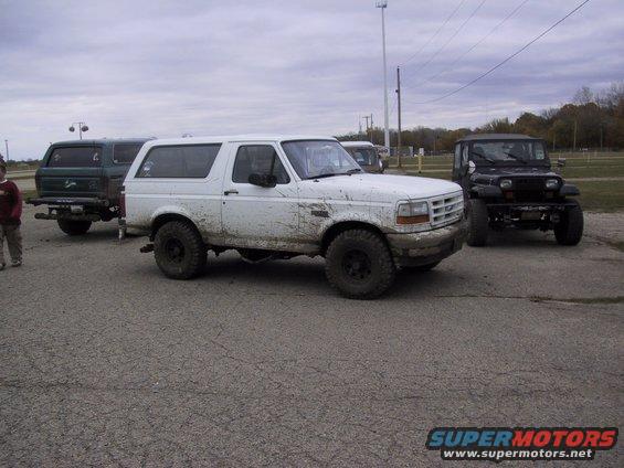 White Ford Bronco covered in mud, parked on asphalt with other vehicles