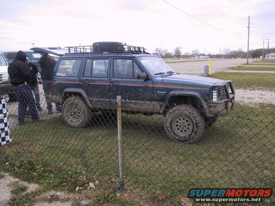 Muddy dark green Jeep Cherokee XJ with roof rack and spare tire, parked on grass near a fence