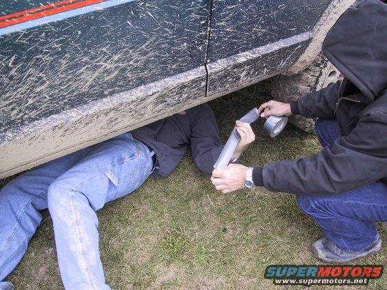 Mud-splattered dark green SUV side with two people working underneath on exhaust pipe with duct tape