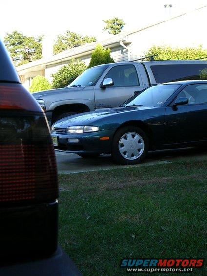 Dark green coupe with silver hubcaps, 3q-front view, parked next to a silver pickup truck
