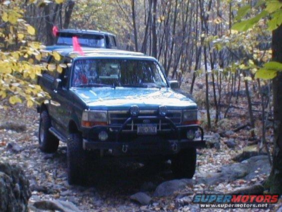 Green Ford Bronco driving on a rocky trail in the woods, another Bronco behind it