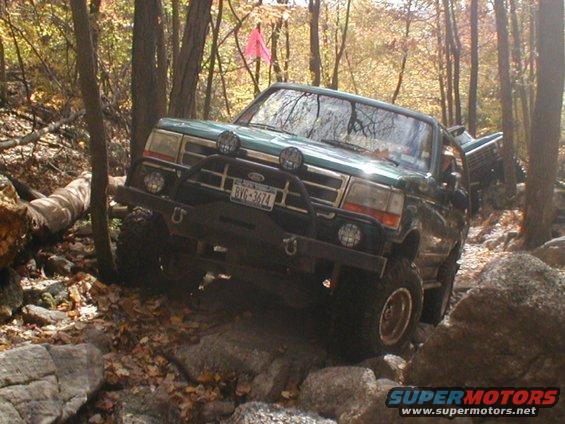 Green Ford Bronco navigating a rocky, wooded trail. Front bumper has auxiliary lights and tow hooks. License plate BYG-3674 is visible