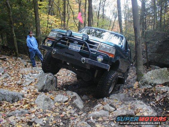 Green Ford Bronco navigating a rocky forest trail, front bumper and headlights visible