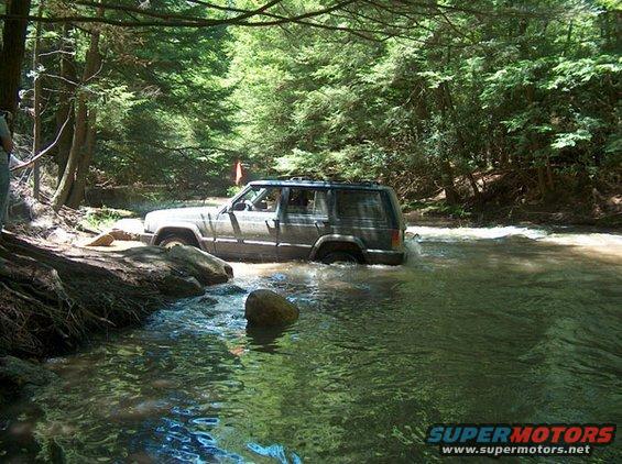 Gray Jeep Cherokee driving through a shallow river in a forest