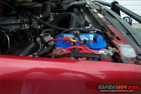 Engine bay showing a blue Optima battery with red cables and various hoses and engine components