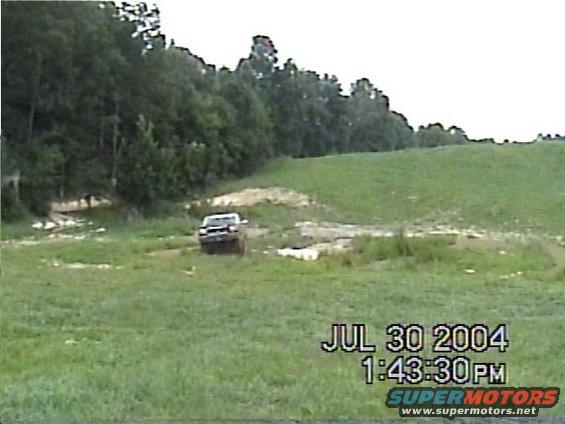 Truck driving through a muddy, grassy field with trees in the background. Timestamp overlay reads JUL 30 2004 1:43:30 PM