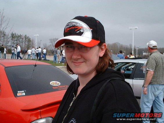 Woman in a black hoodie and Pontiac Racing hat, with a red car and a silver car in the background