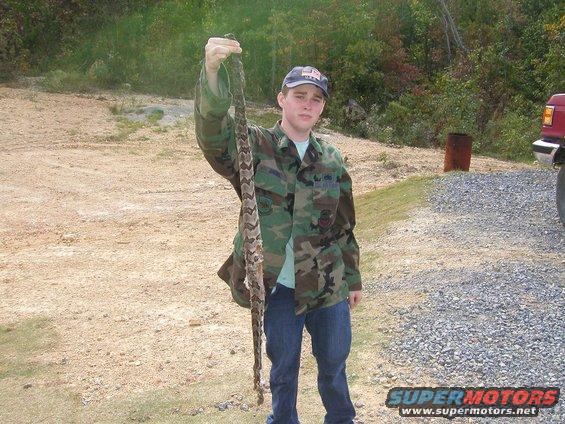 Guy in camo jacket and jeans holding up a long snake