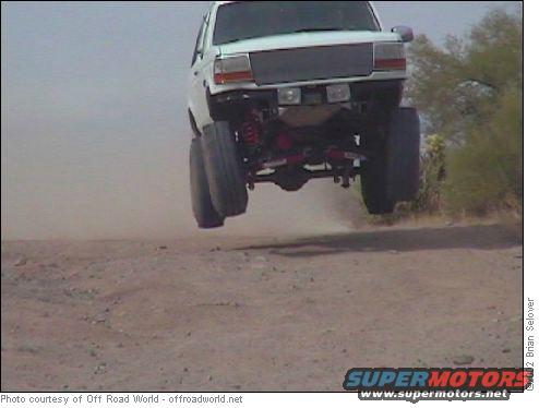 White SUV mid-air over a dirt track, front view, lifted suspension, large tires
