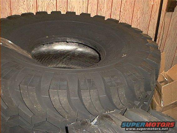 Big off-road tire with aggressive tread pattern, stacked on other tires. Cardboard boxes in background