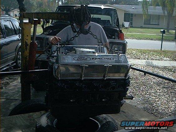 Ford Racing engine with chrome valve covers, suspended by chains. A person is operating the hoist. A black truck and a silver SUV are visible in the background