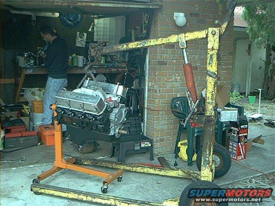 Engine on an orange stand in a garage, with a yellow engine hoist over it. A person is visible in the background