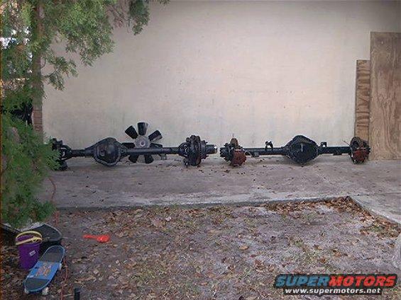 Two vehicle axles are laid out on the ground in front of a wall. One has a fan attached