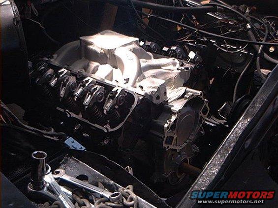 Engine block with cylinder heads exposed, intake manifold removed, showing valve train components. Tools and chains are in the foreground