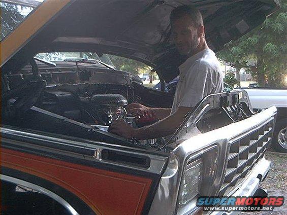 Man working on the engine of a 1970s Ford truck. Another vehicle is visible in the background