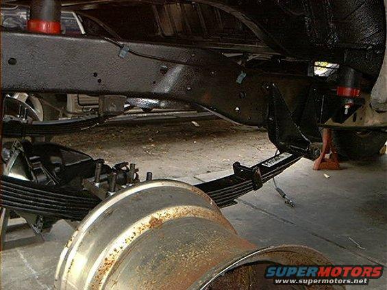 Underside of a vehicle showing the frame, leaf springs, and part of a wheel drum. Some rust is visible on the drum