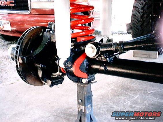 Close-up of a black axle with red coil springs and white shock absorbers, supported by jack stands. A red vehicle with a GMC logo is partially visible