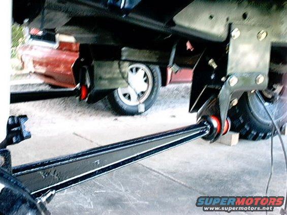 Close-up of a black metal suspension arm with red bushings, attached to a vehicle frame. Another vehicle is partially visible in the background