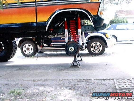 Orange and black Ford Bronco lifted on jack stands, showing suspension modifications, with a white sedan underneath