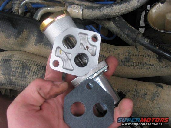 Hand holding a metal engine part with a gasket against a background of hoses and engine components