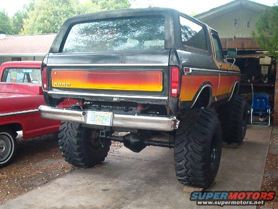 Rear view of a lifted 1979 Ford Bronco with orange and yellow stripe paint. Another red truck is parked next to it