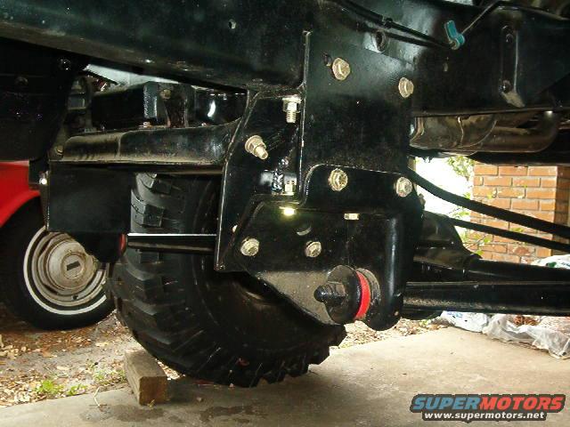 Black suspension bracket with red bushing attached to a large tire. Another vehicle's wheel is visible