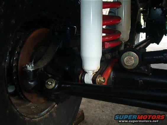close-up of a vehicle's front suspension and steering linkage, showing a white shock absorber with a red coil spring, brake rotor, and black tie rod