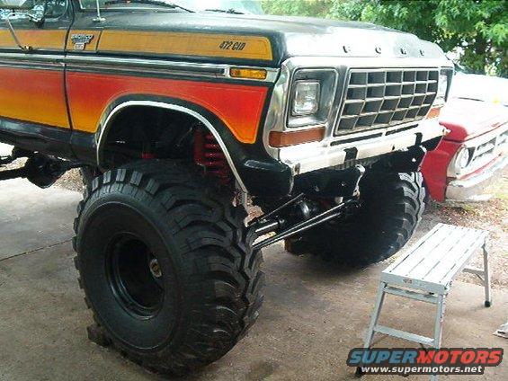 Lifted 1979 Ford Bronco with orange and red gradient paint, large off-road tires, and "472 CID" badge. Another red truck is visible behind it