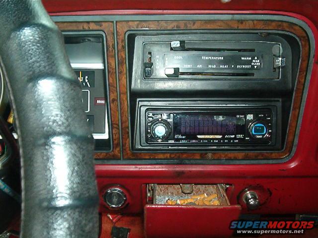 Car dashboard with wood trim, climate controls, aftermarket stereo, and open ashtray filled with cigarette butts
