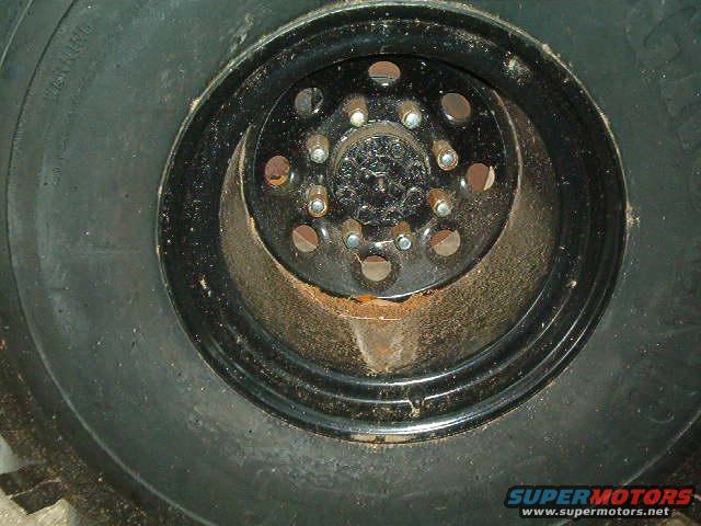 Close-up of a black wheel with a large tire. The wheel has multiple lug nuts and circular cutouts. Some rust is visible on the wheel's inner rim