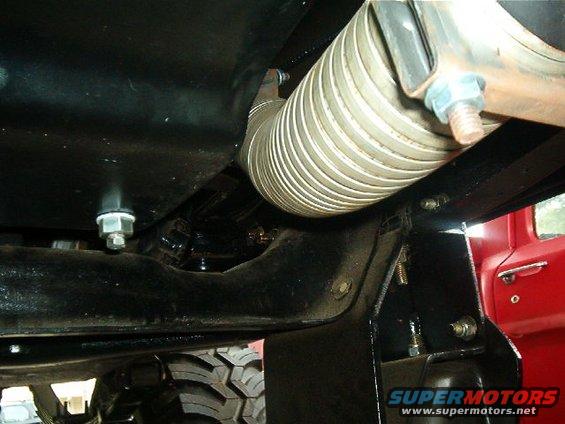 Underside view of a vehicle's exhaust pipe and chassis components. Tire tread visible