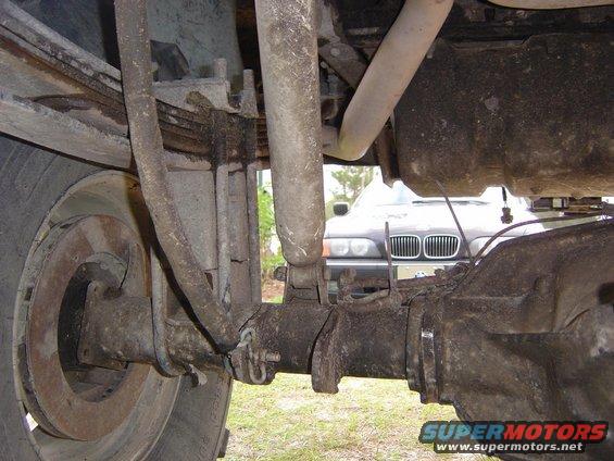 Close-up of a vehicle's rear axle, leaf springs, and shock absorber. Another car is visible in the background