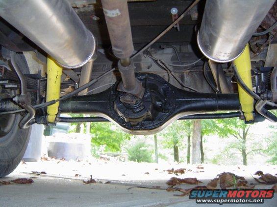 Underside view of a vehicle's rear axle, driveshaft, and dual exhaust pipes with yellow shock absorbers