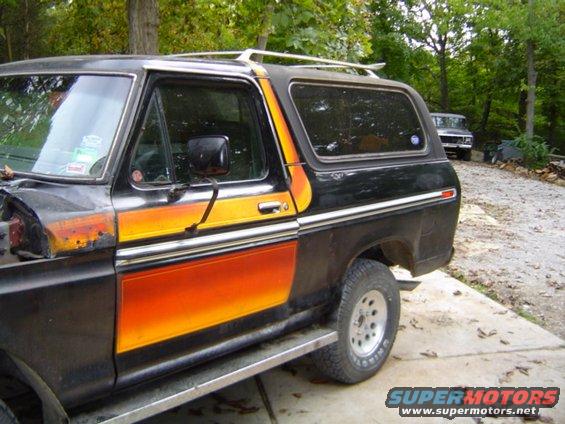 Black 1978 Ford Bronco with orange and yellow stripe graphics, parked on a concrete driveway. Another vehicle is visible in the background