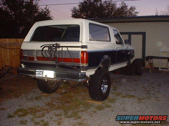 White and blue Ford Bronco with a rear spare tire carrier, lifted suspension, and chrome wheels