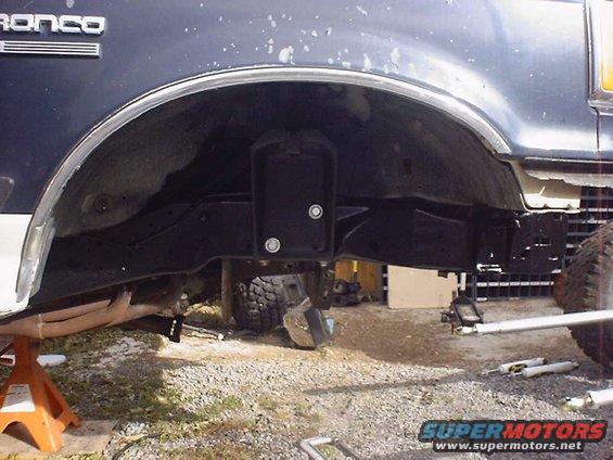 Bronco emblem on dark blue vehicle body, showing suspension and frame parts underneath. Vehicle is on jack stands
