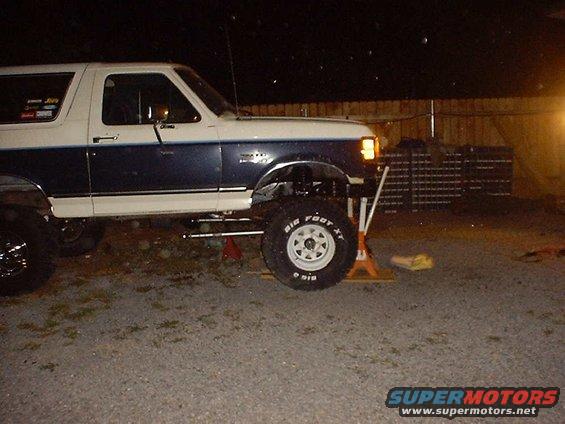 Side view of a lifted Ford Bronco with "Bigfoot XT" tires, supported by jack stands at night