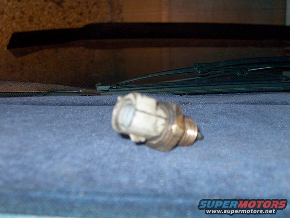 Brass automotive sensor with white plastic connector, resting on blue carpet near windshield wipers
