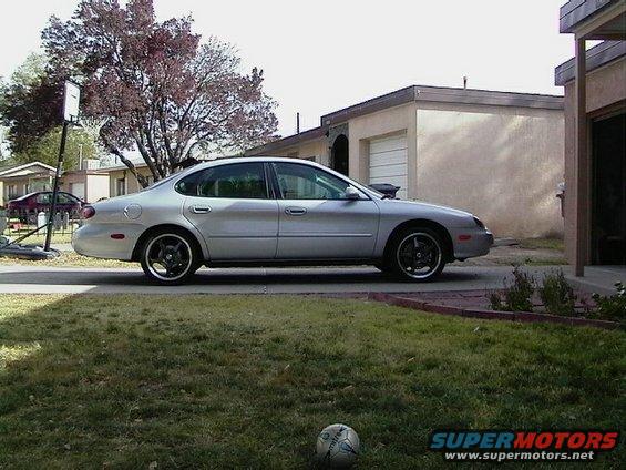 Silver sedan parked in a driveway, side view. Aftermarket black wheels, slight damage on front fender