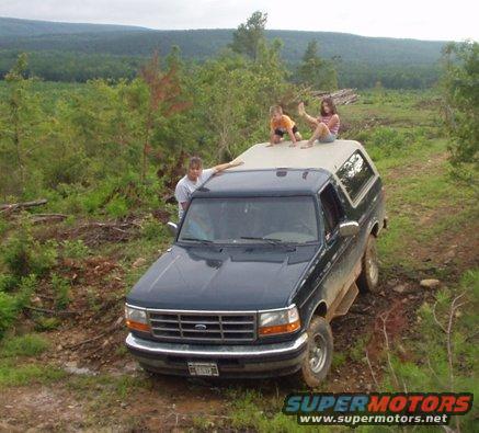 Dark blue Ford Bronco parked on a dirt road with two kids on the roof and one person leaning on the side