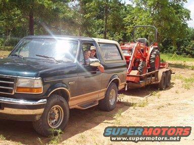 1995 Ford Bronco towing a red tractor on a dual-axle trailer on a dirt road