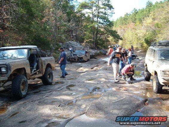 Several off-road vehicles navigating a rocky, wet trail with trees in the background