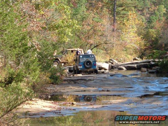 Two Jeeps driving on a rocky stream bed with fall foliage in the background