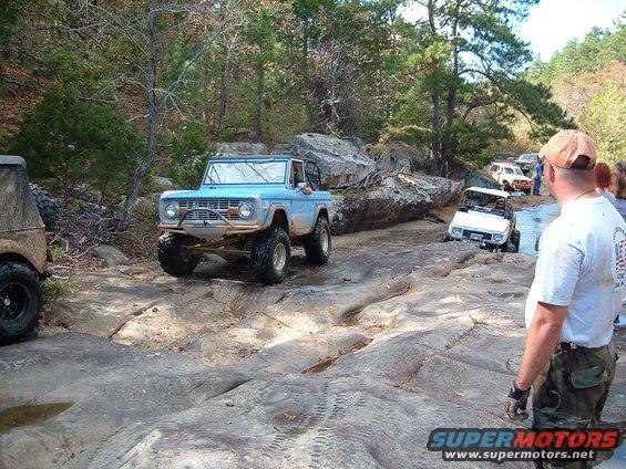 Blue first-gen Ford Bronco and other off-road vehicles navigating a rocky, wet trail