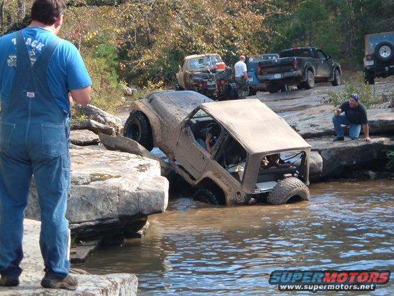 Muddy off-road vehicles crossing a rocky stream, one vehicle is partially submerged